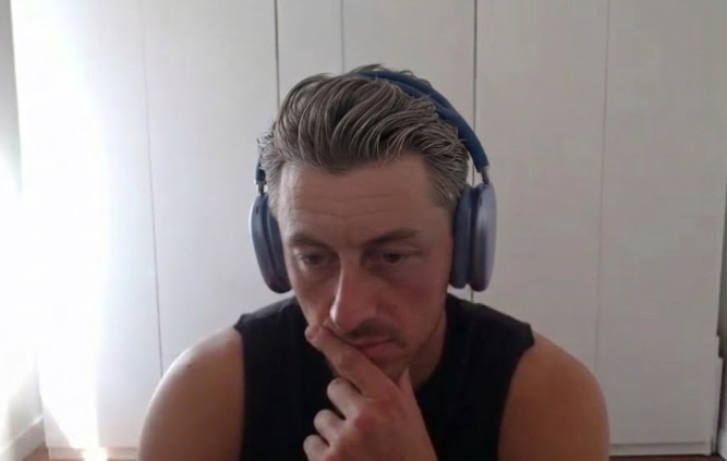 A man with slicked-back hair wearing a black sleeveless shirt and blue over-ear headphones, looking down thoughtfully with his hand near his chin in front of white cabinets.