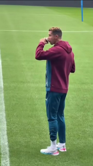 A young man wearing a maroon hoodie and teal training pants drinks from a cup while standing on a grassy field, likely a soccer training ground.