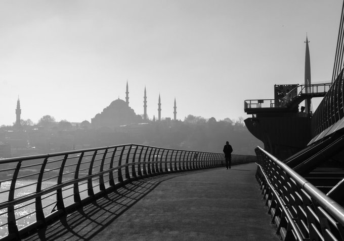 A black-and-white photograph features a person walking along a waterfront path with railings, with the silhouette of a large mosque with multiple minarets visible in the misty distance.