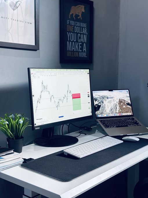 A minimalist home office desk setup featuring a Lenovo monitor displaying a trading chart, a laptop, a keyboard, and a motivational sign reading 'IF YOU CAN MAKE ONE DOLLAR, YOU CAN MAKE A MILLION MORE.'