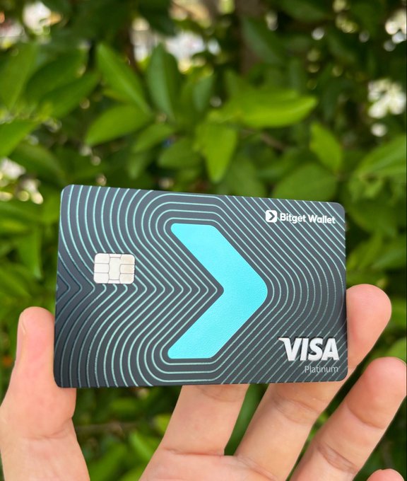 A hand holds a dark gray Bitget Wallet Visa Platinum card with a teal arrow design and EMV chip, set against a backdrop of green leaves. The image aligns with the "GM @BitgetWalletCN" post by showcasing the brand's physical Visa Platinum card product.