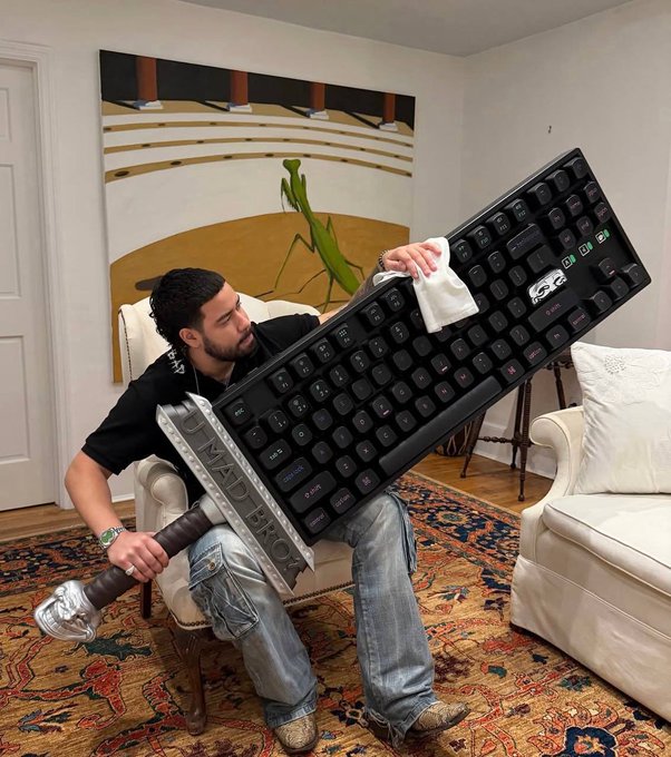 A young man sits in a chair holding a comically oversized prop sword with the word 'EXCALIBUR' written on the blade, while also holding a massive, oversized black computer keyboard that is nearly as tall as he is.