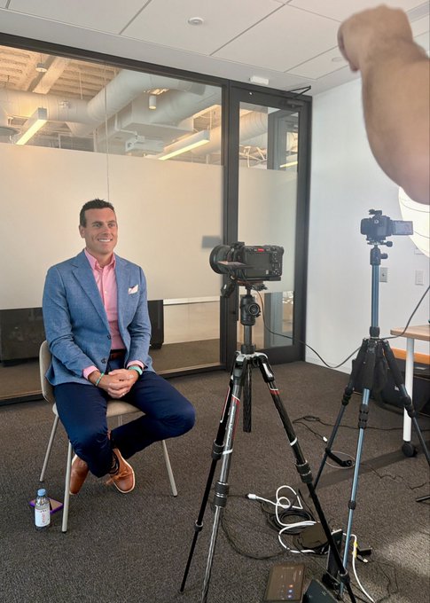 A man in a blue blazer and pink shirt being filmed in an office setting, seated across from two cameras on tripods with a hand visible directing in the foreground.