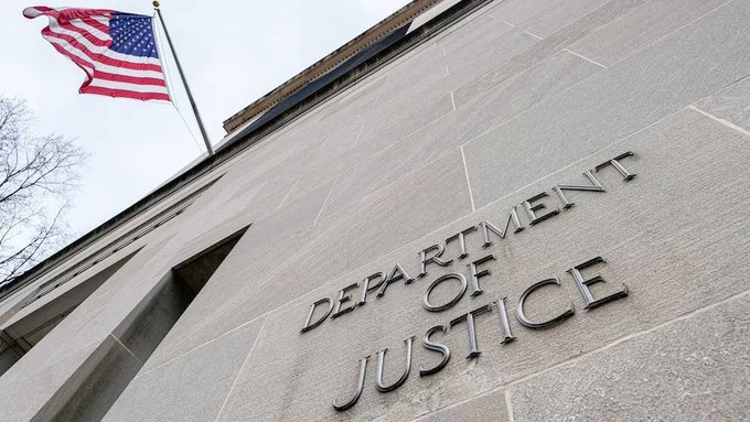 A low-angle photograph of the Department of Justice building exterior, showing the carved stone lettering 'DEPARTMENT OF JUSTICE' on the building's facade with an American flag waving on top against a cloudy sky.