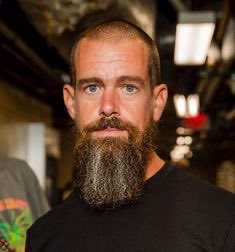 The image features a man with a beard wearing a black shirt, standing indoors. The background of the photo shows an indoor setting with a ceiling that appears to be made of metal or steel. There are also other people present in the scene, but they are not the main focus of the image. The post about Jack Dorsey's Cash App has been mentioned multiple times and is related to the image as it features a man wearing a black shirt and a beard.