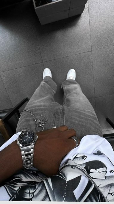 A first-person perspective photo looking down at grey jeans, white sneakers, and a dark-skinned hand wearing a silver Rolex watch and rings, with a silver chain and pendant visible.