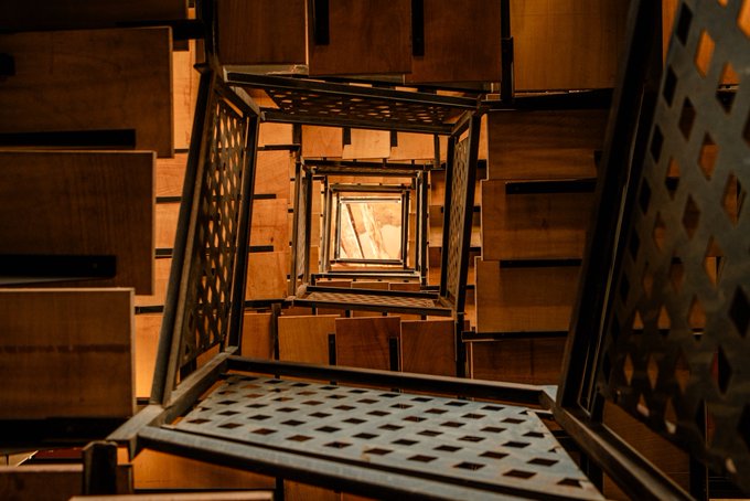 The image shows a spiral staircase made of metal bars that are arranged in a way that they appear to be going up or down. The staircase has a unique design, with each step having a grid-like pattern. The stairs are located inside a room, which is dimly lit and filled with wooden boxes stacked on top of one another.