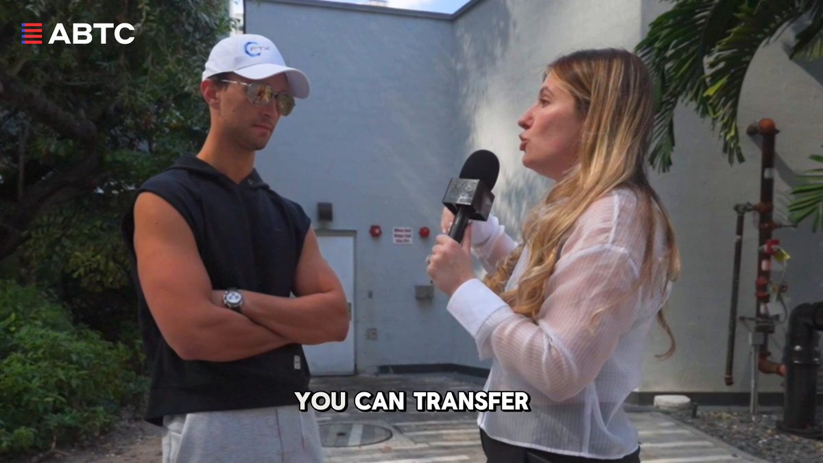 The image shows two people, one wearing a black shirt and the other wearing a white shirt, standing next to each other with their arms crossed. They are engaged in conversation or having an interview, as indicated by the text "You can transfer". The background of the image is blurred, but it appears to be outdoors, possibly near some trees.