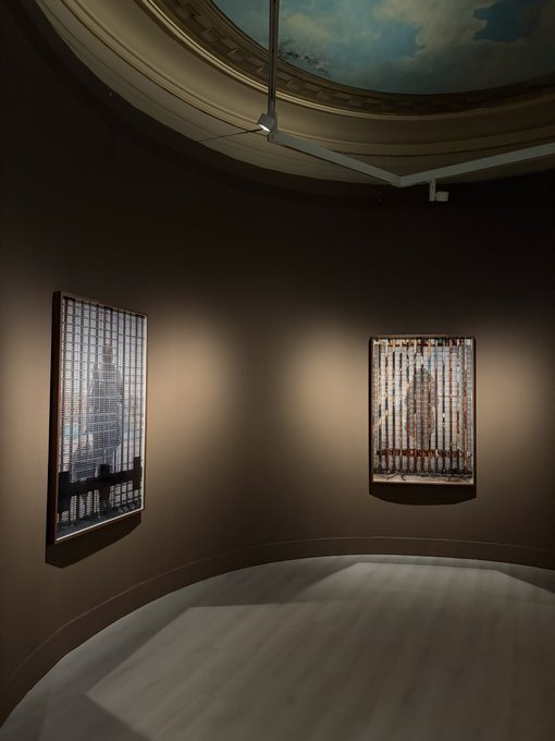 A dimly lit art gallery room with curved brown walls displaying two framed artworks, featuring a painted sky ceiling and a marble floor.