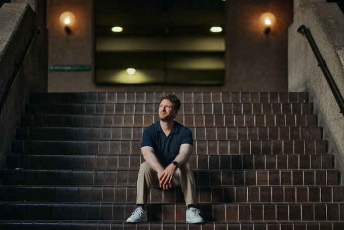 A young man sits contemplatively on a wide brick staircase in a dimly lit corridor, hands clasped and gaze directed off to the side, as if thoughtfully reflecting on a new chapter. The moody, cinematic composition aligns with the welcoming post "Welcome to the team Martin!", framing him as someone stepping into a significant new role.