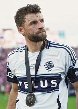 Thomas Müller is pictured wearing his Vancouver Whitecaps FC jersey with a medal around his neck, a club he joined after his legendary Bayern Munich career—the same club that rejected a €100 million Manchester United bid for him in 2015. The image likely reflects his post-Bayern chapter, contrasting the career move that ultimately did happen with the Premier League transfer that never materialized.