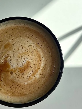 An overhead close-up view of a dark cup filled with a frothy, light brown coffee beverage sitting on a white surface.