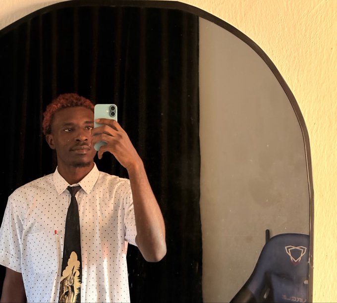 A mirror selfie of a young man with reddish-brown hair wearing a white polka dot shirt and a black tie with a design, holding a light blue phone to take the photo, with a gaming chair visible in the background.