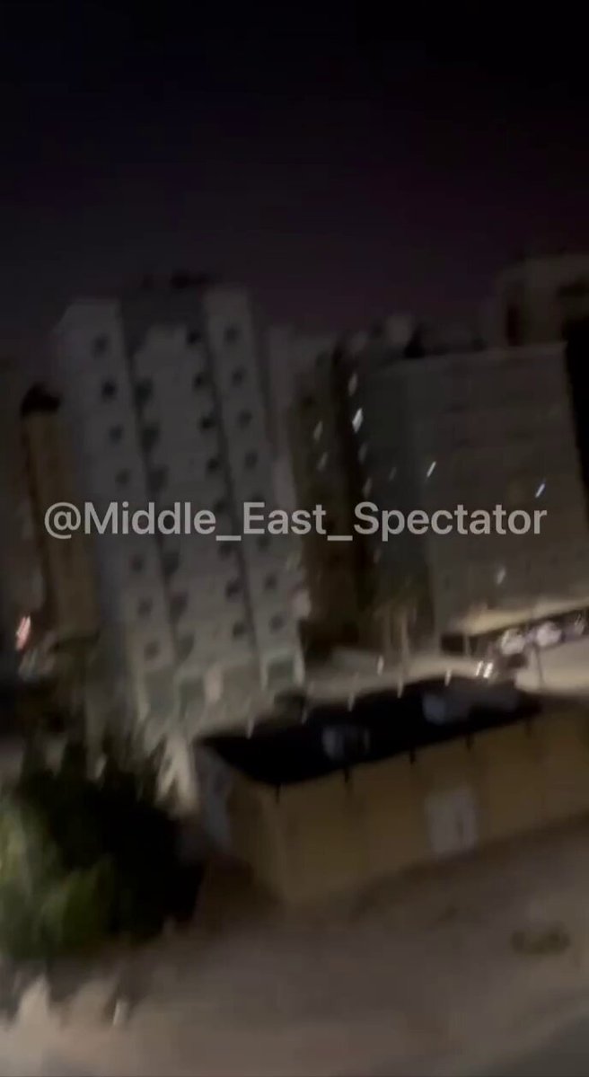 The image shows a city street at night with tall buildings, including an apartment building. The sky above appears dark, creating a contrast between the illuminated buildings and their surroundings. There are also cars parked along the street, adding to the urban atmosphere of the scene.