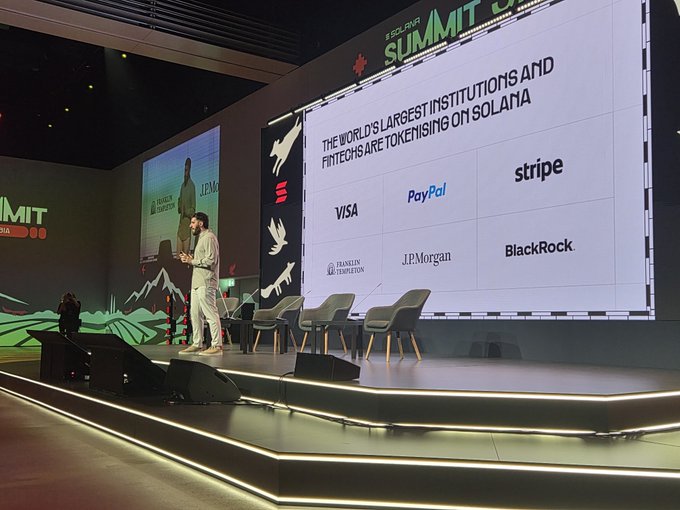 A speaker stands on stage at the Solana Summit, with a large screen behind him displaying 'THE WORLD'S LARGEST INSTITUTIONS AND FINTECHS ARE TOKENISING ON SOLANA' alongside logos of VISA, PayPal, stripe, Franklin Templeton, J.P.Morgan, and BlackRock.
