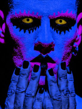 A person with vibrant blue and pink face paint, yellow eyes, and black chipped nail polish holds their hands up to their face in a dramatic pose. The chaotic, vivid aesthetic of the image mirrors the eccentric, crypto-fueled plea for funding a car purchase with Tezos (XTZ) in exchange for art.