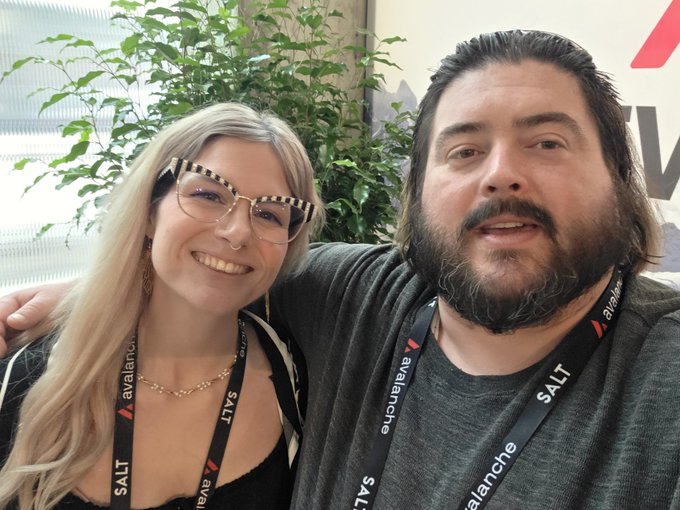 A blonde woman wearing checkered glasses smiles next to a bearded man; both are wearing lanyards branded with 'SALT' and 'Avalanche'.