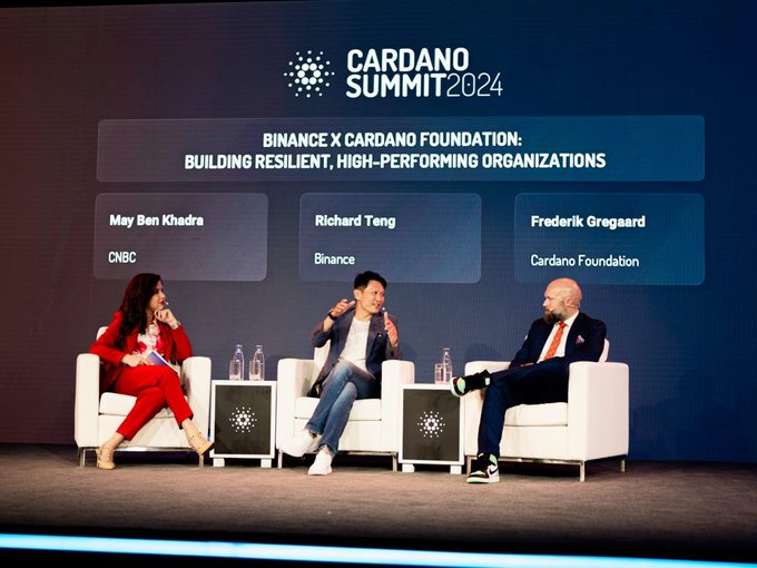 The image shows three people sitting on chairs around a table, with a banner behind them that reads "CARDANO Summit 2014". The individuals are engaged in conversation, possibly discussing the event or its content. One person appears to be wearing a red shirt while another is dressed in black and white. A third person can be seen seated at the end of the table.