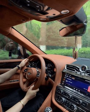 A view from the driver's seat of a luxury car featuring a tan leather interior, showing hands on the steering wheel with an infotainment screen displaying "107.4 MHz" and trees visible through the windshield, contrasting the opulent lifestyle implied by the post about a crypto trader's success and multiple properties.