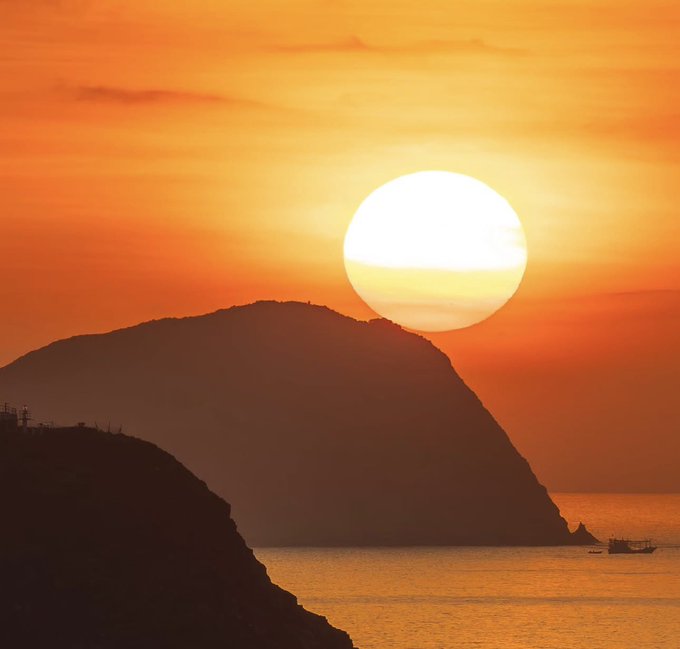 A stunning sunset over a coastal landscape with a large silhouetted mountain or hill in the foreground and the bright sun positioned just above it, casting an orange glow across the water and sky.