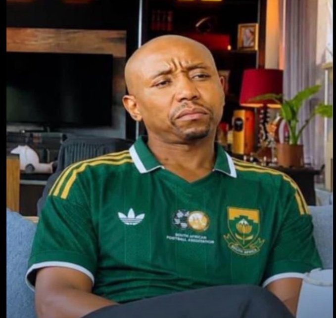 A man with a shaved head and skeptical expression sits on a gray couch wearing a green Adidas jersey with the South African Football Association logo.