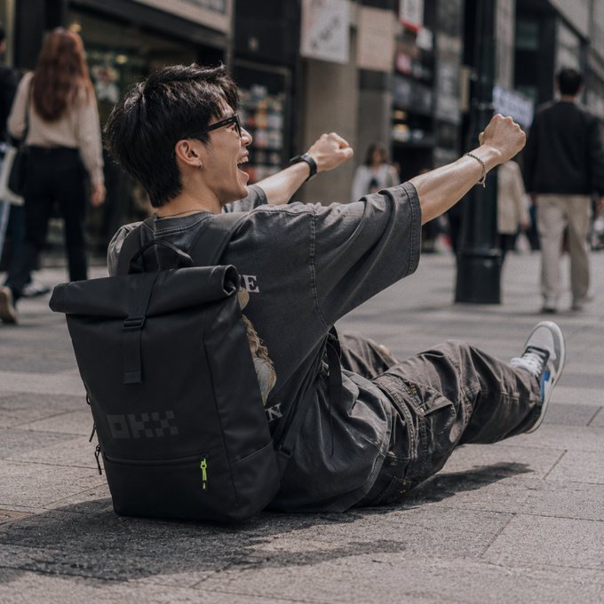 A young man wearing glasses, a dark shirt, and a DKX backpack sits on the pavement with arms outstretched in celebration.