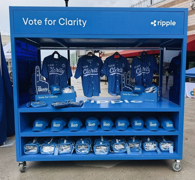 The image shows a blue display case filled with various baseball caps, likely for sale at an event or store. There are multiple rows of hats displayed inside the case, showcasing different styles and colors. The hats appear to be organized by size and style, making them easily accessible for customers. The display is situated on wheels, allowing it to be moved around as needed. The caption "VOTE FOR CLARITY @Ripple" suggests that this event or store may have a connection to the Ripple cryptocurrency, possibly promoting both baseball caps and digital currency awareness.