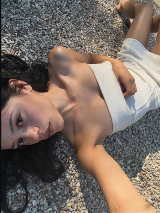 A selfie of a woman lying on gravel in the sun, wrapped in a white towel.
