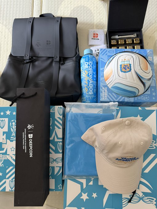 A collection of official Argentina national team merchandise from Samsung, including a black backpack, a soccer ball, a blue "Copa del Mundo" water bottle, a beige "Vamos Argentina" cap, a blue shirt, a "DEEPCOIN" branded gift bag, and a black box with gold tokens, all displayed alongside an "AFA" branded gift box. The items connect to the post's mention of the upcoming Argentina vs England match, sponsored by partners Deepcoin and Evie Crypto, with the merchandise serving as promotional gifts for fans or influencers.