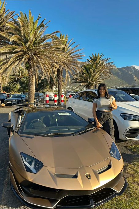 A woman poses next to a matte gold Lamborghini Aventador SVJ in a scenic location with palm trees and mountains in the background, embodying the luxurious lifestyle vibe of the $River points promotion shared in her post.