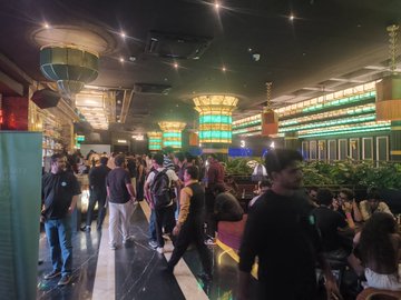 A crowded indoor venue resembling a casino or poker room, with many people standing among tables while large screens display green graphics and betting information in the background.