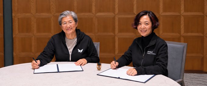 Two women sit at a round table in a formal setting, each holding a pen over signed documents, with one wearing an AMD jacket, suggesting a partnership or signing event.