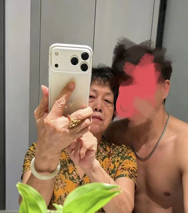A mirror selfie of an older woman in a floral shirt taking a photo with an iPhone while a shirtless man with a pink/red censor bar over his face stands behind her.