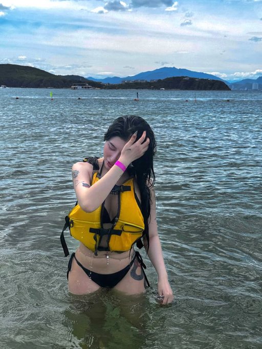 A woman in a yellow life jacket and black bikini stands in shallow ocean water, brushing her wet hair back from her face with a contemplative expression against a backdrop of distant mountains and cloudy skies. The caption's theme of missed profits connects to her reflective, wistful posture—standing in the water as if she's been "drowning" in regret over unrealized gains, with the vast sea visually emphasizing the depth of her financial losses.