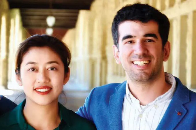A smiling young woman with dark hair and red lipstick wearing a green top stands next to a smiling young man with dark hair wearing a blue blazer and white shirt in a hallway with arches.