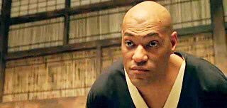 A close-up of a bald man in traditional Japanese clothing with an intense, serious expression in what appears to be a scene from a martial arts film.