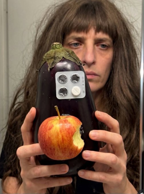 A woman holds an eggplant that has been creatively decorated to look like a smartphone (complete with camera lenses and a pill-shaped button), with a bitten apple attached to it as a logo, humorously recreating a phone design.