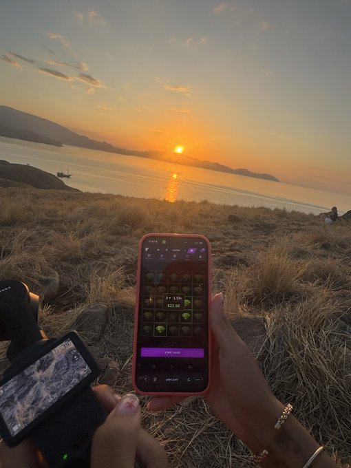 A first-person view captures hands holding a smartphone displaying a purple-themed app showing "3.13s" and "$22.50" with a "Start Earning" button, alongside a camera with a flip-out screen, all set against a stunning sunset over a tranquil lake and distant mountains—perfectly illustrating the "trekking, vlog and yeeting in sunset" adventure.