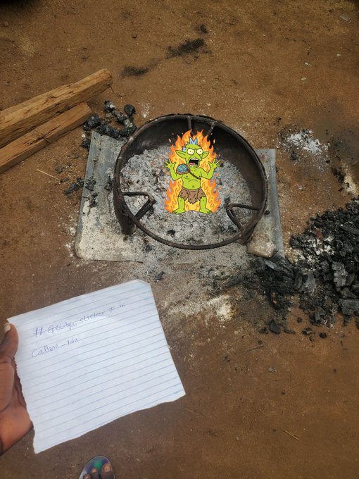 A hand holds a note addressed to "Catliano" near a charcoal grill containing a cartoon zombie sticker on the ashes, with scattered charcoal and wood nearby.