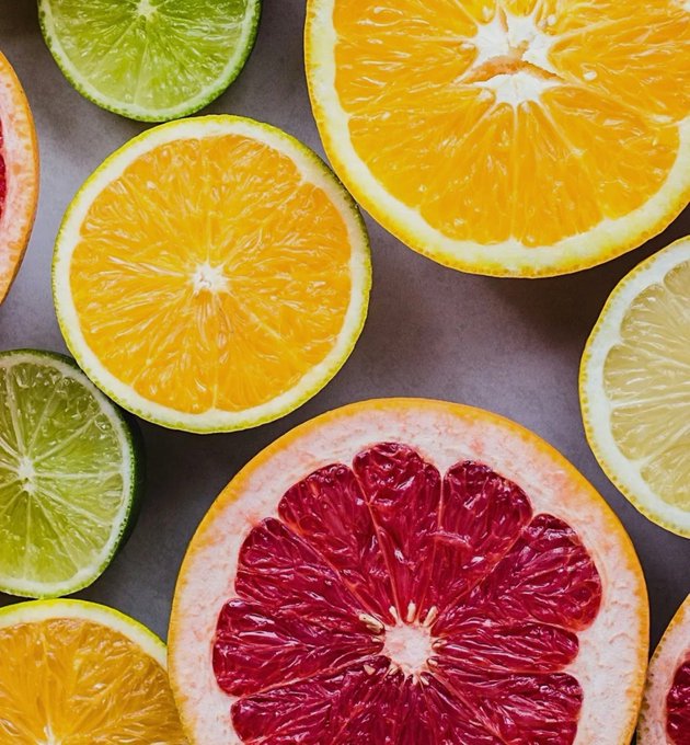 A vibrant overhead photograph of various sliced citrus fruits including oranges, limes, lemons, and a blood grapefruit arranged on a dark surface.