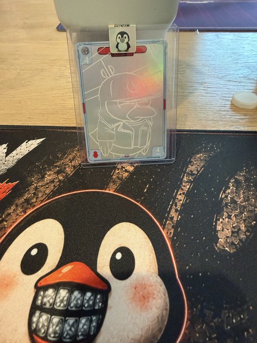 A holographic card displayed in a clear stand, featuring an engraved illustration of a chubby character holding a mug, sitting on a dark mat with a cartoon face design and clipped with a penguin logo.