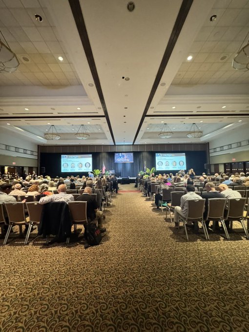 A large conference room filled with attendees seated in rows of chairs, facing a stage with multiple presentation screens at the front.