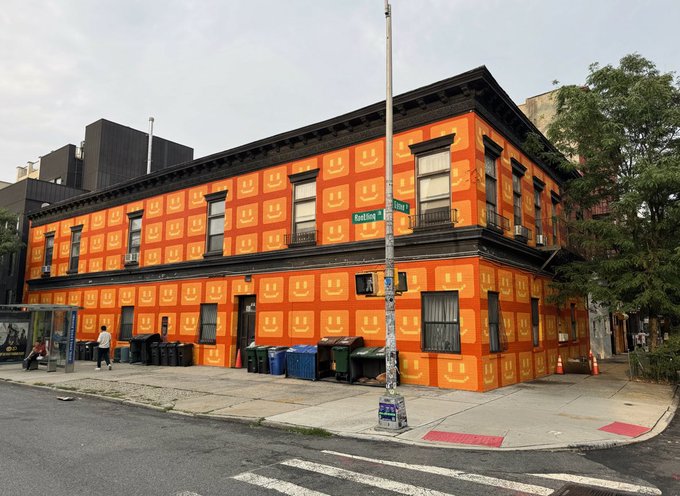 An urban street corner featuring a two-story building covered in bright orange wrapping printed with a smiley face logo, located at the intersection of Knickerbocker Ave.