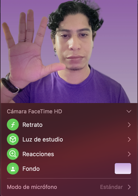A man holds his hand up toward the camera in front of a purple background, with a FaceTime HD camera settings menu (Retrato, Luz de estudio, Reacciones, Fondo) visible below him.