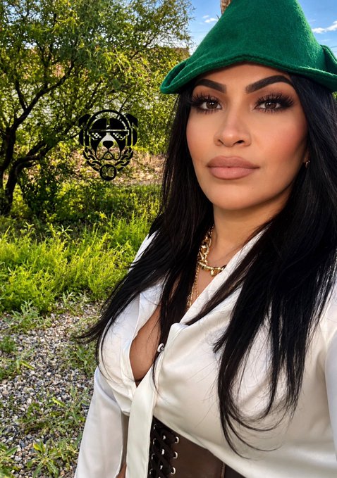 A woman with long dark hair wearing a green hat, white satin blouse, and gold chain poses outdoors with a decorative skull logo overlaid.