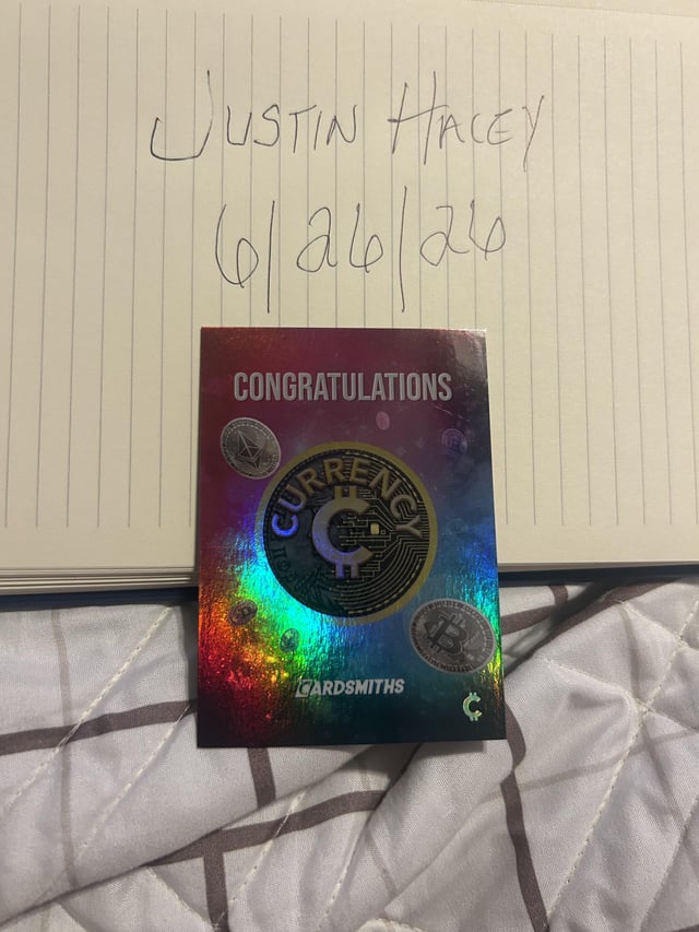A holographic 'CONGRATULATIONS' card featuring 'CURRENCY' and 'CARDSMITHS' branding, placed on a notebook with 'Justin Hacey 6/26/26' handwritten above it.