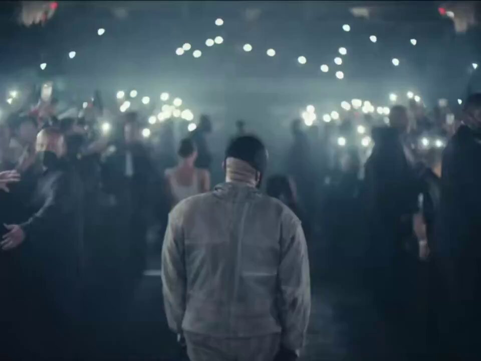 The image shows a man standing in front of a crowd, with many people surrounding him. The man appears to be wearing a grey jacket and has his back turned towards the camera. In the background, there are numerous cell phones held up by individuals, suggesting that they might be taking photos or videos during this event. The overall atmosphere seems lively and energetic as everyone is engaged in capturing memories of the moment.