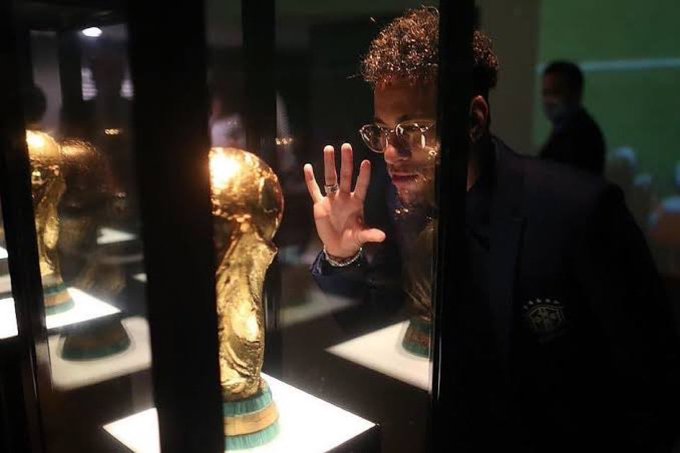 A young man with curly hair and glasses in a dark blazer looks through a glass display case at a golden trophy on an illuminated pedestal in a museum-like setting.