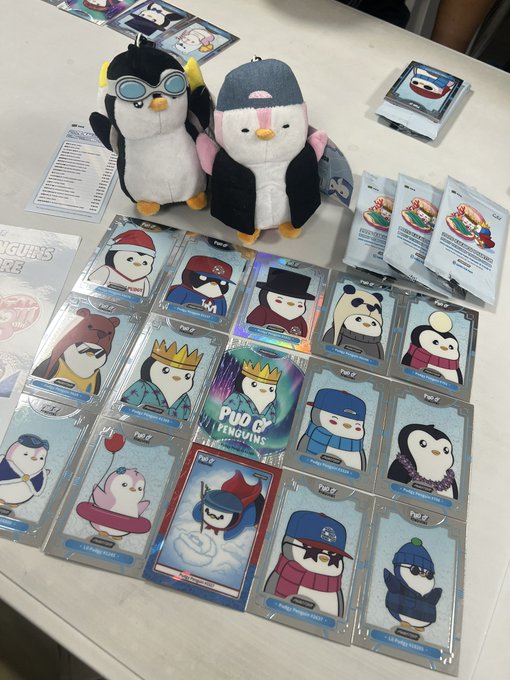 A display of 'Pudgy Penguins' trading cards and plush toys on a white table, featuring various penguin characters in different outfits and designs still in their protective sleeves.