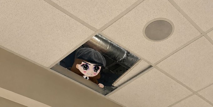 A photo showing a large anime character cutout or poster of a girl with a beret peeking out from an open ceiling tile in a drop-ceiling, revealing the ductwork above.